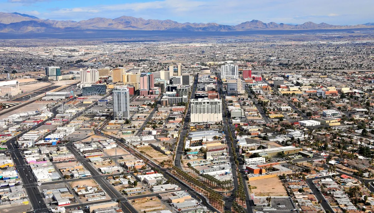 Aerial view of downtown Las Vegas with tall buildings and a long, straight road, with mountains in the background.